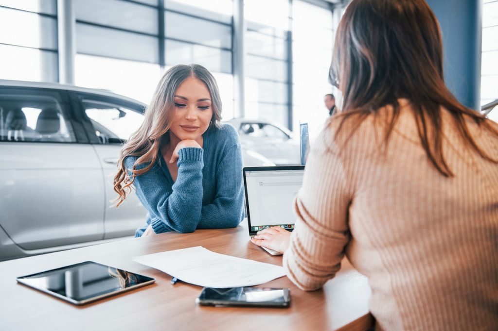 Cheerful woman sits with assistant in car shop and buying new vehicle.